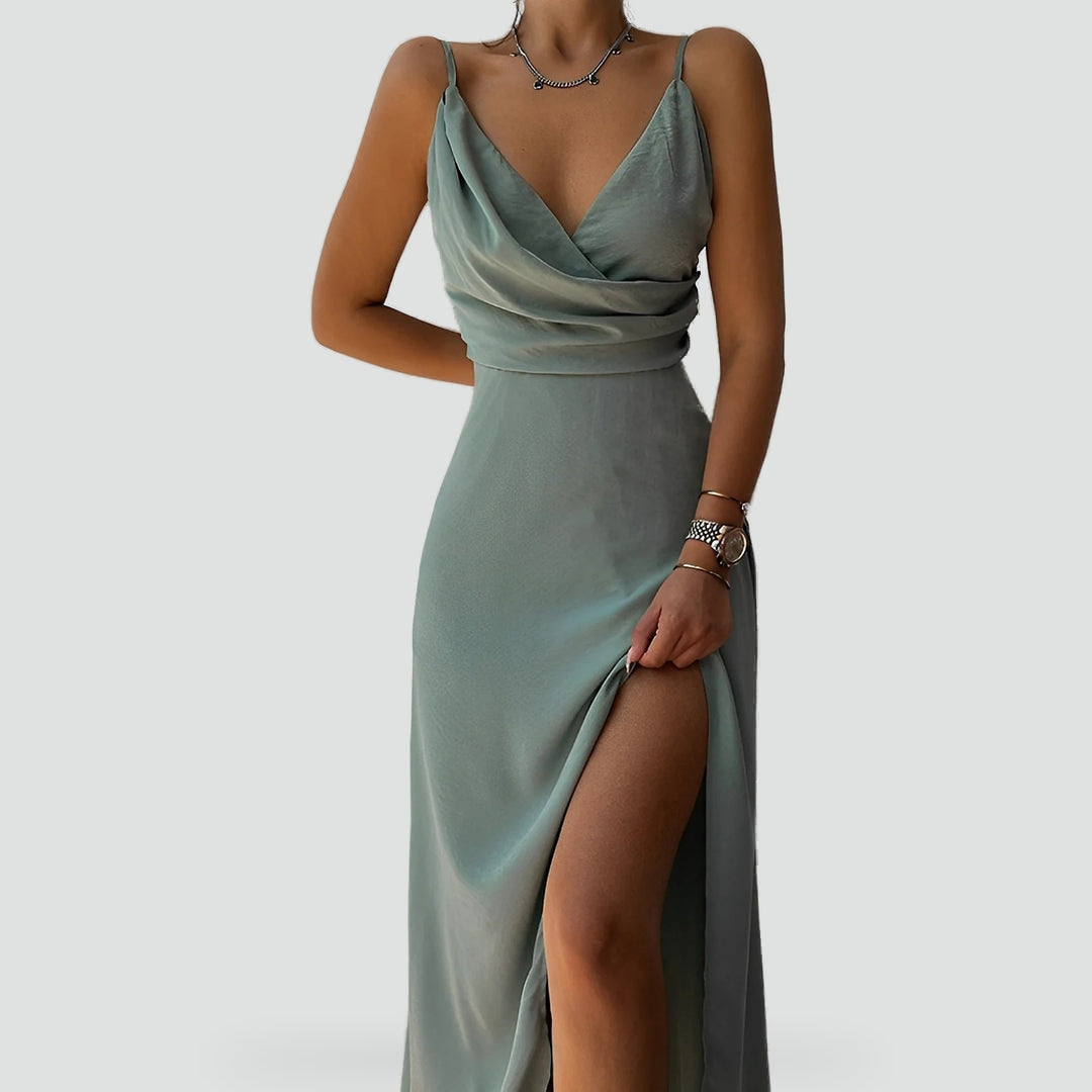 Femme portant une élégante robe longue fluide de couleur vert sauge, à fines bretelles et décolleté drapé. La tenue présente une fente haute sur la jambe, ajoutant une touche sensuelle et sophistiquée. Le tissu satiné reflète délicatement la lumière, créant un effet doux et luxueux, parfait pour une soirée ou un événement chic.